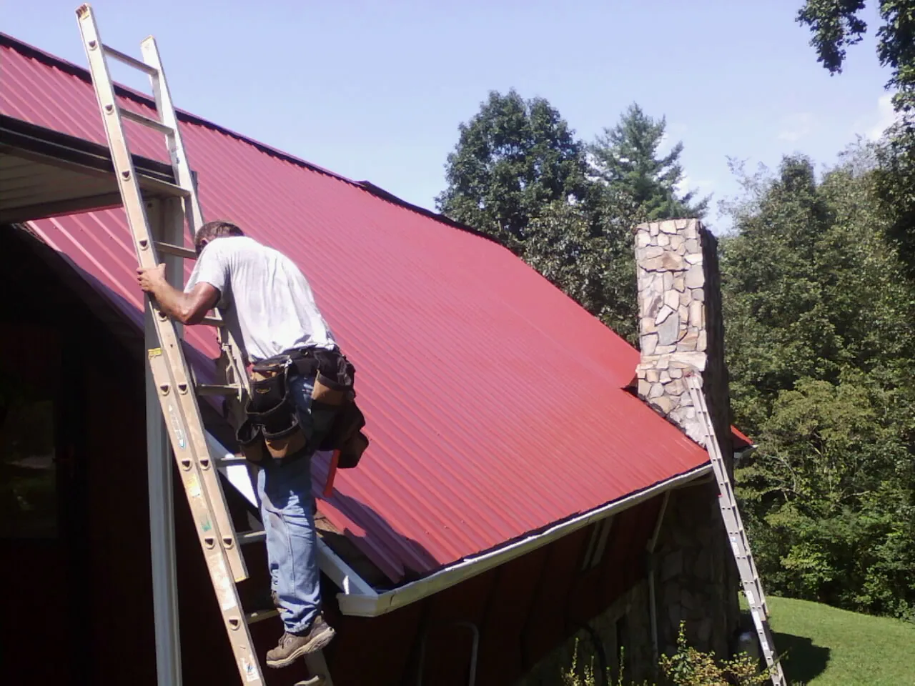 Roofer on a ladder beside a red metal roof and stone chimney — Soffit & Fascia Repair by Roofing Brothers in South Bethlehem